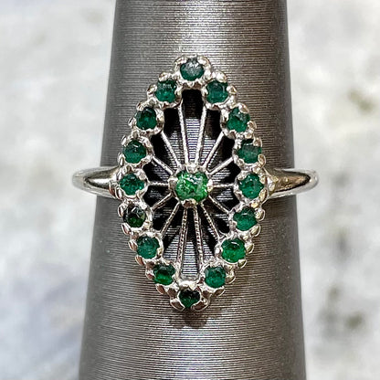 A vintage marquise shape white gold ring set with a halo of round cut emeralds around a single center stone.