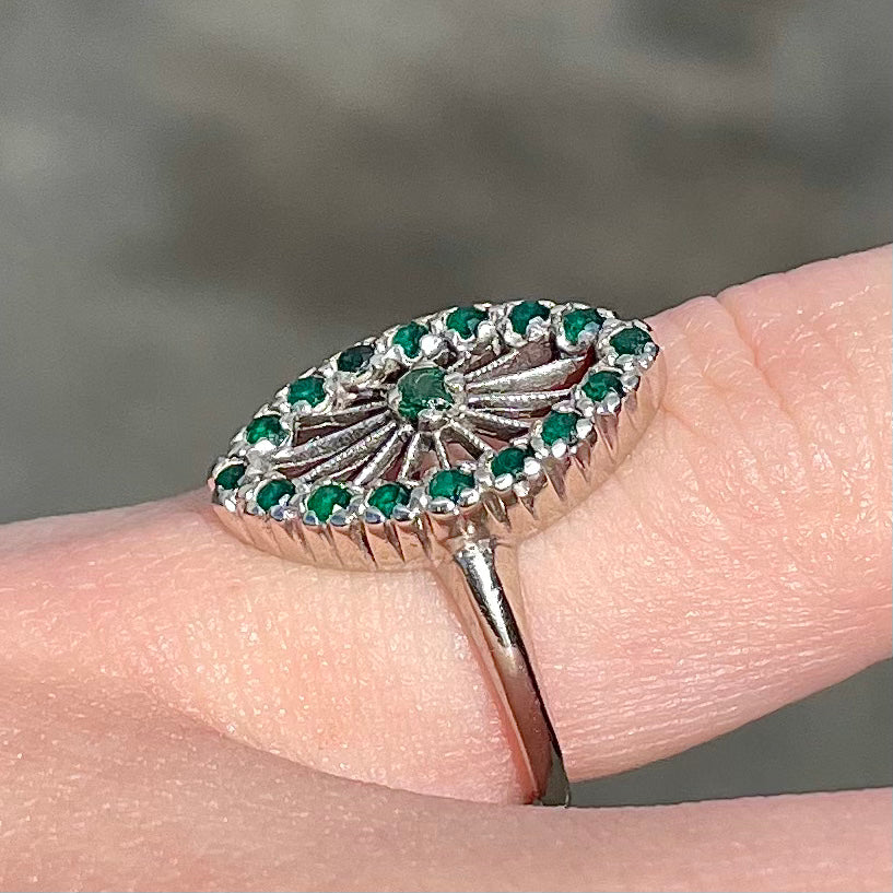 A vintage marquise shape white gold ring set with a halo of round cut emeralds around a single center stone.