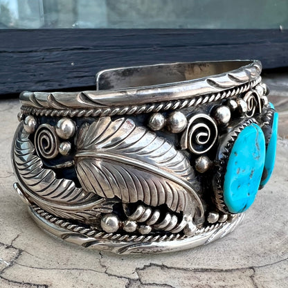 An ornate men's sterling silver turquoise cuff bracelet.  There are three turquoise stones set with silver feathers.