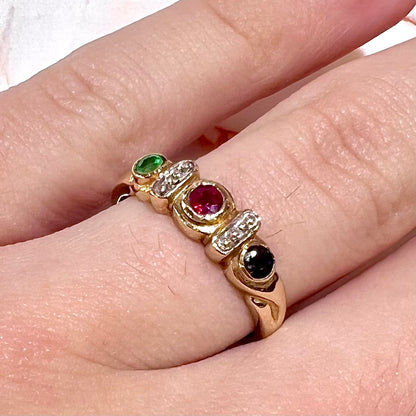 Three stone ring set with blue sapphire, lab created ruby, emerald, and two small diamonds in yellow gold.