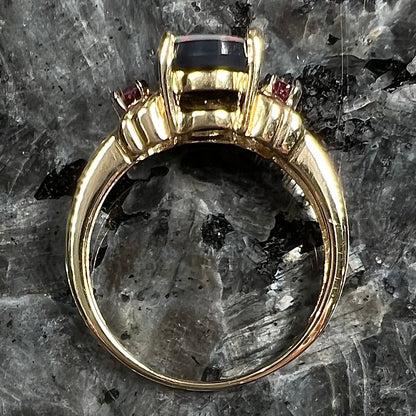 A yellow gold ring prong set with a black opal doublet cabochon and two round ruby accents.