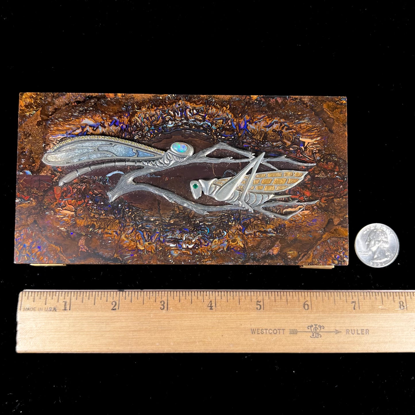 A stone box made from Koroit, Australian boulder opal featuring the motif of a dragonfly and cricket made from sterling silver, set with yellow diamonds, an emerald, and a crystal opal.