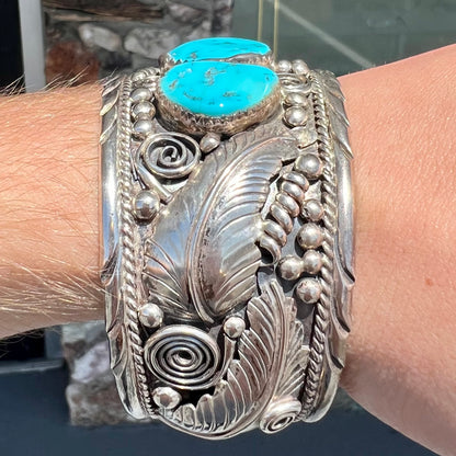 An ornate men's sterling silver turquoise cuff bracelet.  There are three turquoise stones set with silver feathers.