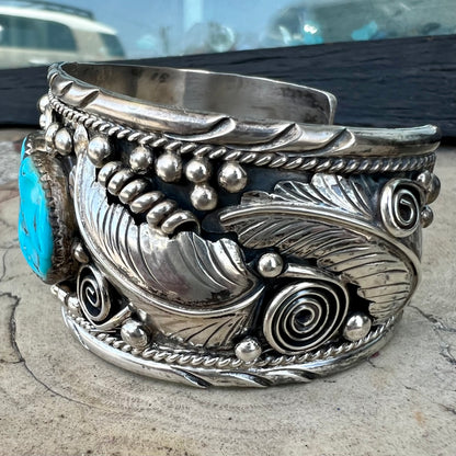 An ornate men's sterling silver turquoise cuff bracelet.  There are three turquoise stones set with silver feathers.