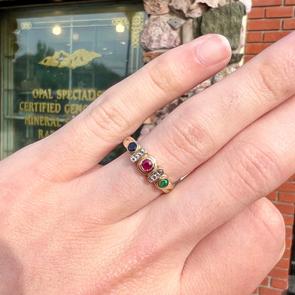 Three stone ring set with blue sapphire, lab created ruby, emerald, and two small diamonds in yellow gold.