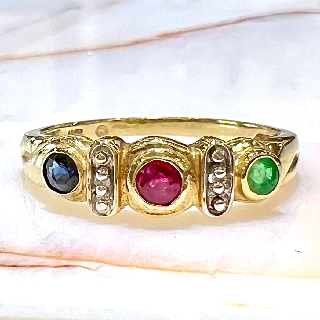 Three stone ring set with blue sapphire, lab created ruby, emerald, and two small diamonds in yellow gold.