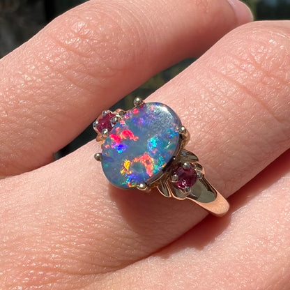 A yellow gold ring prong set with a black opal doublet cabochon and two round ruby accents.