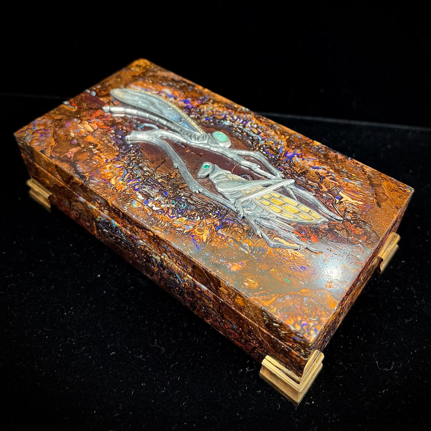 A stone box made from Koroit, Australian boulder opal featuring the motif of a dragonfly and cricket made from sterling silver, set with yellow diamonds, an emerald, and a crystal opal.