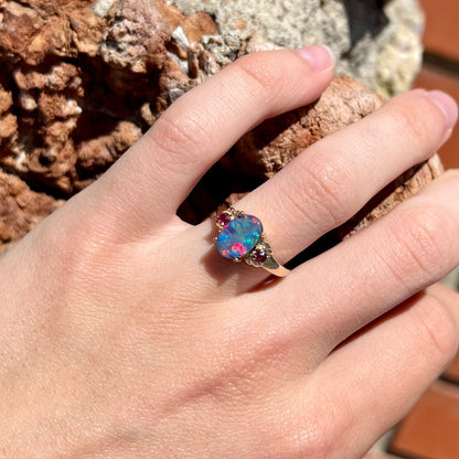 A yellow gold ring prong set with a black opal doublet cabochon and two round ruby accents.