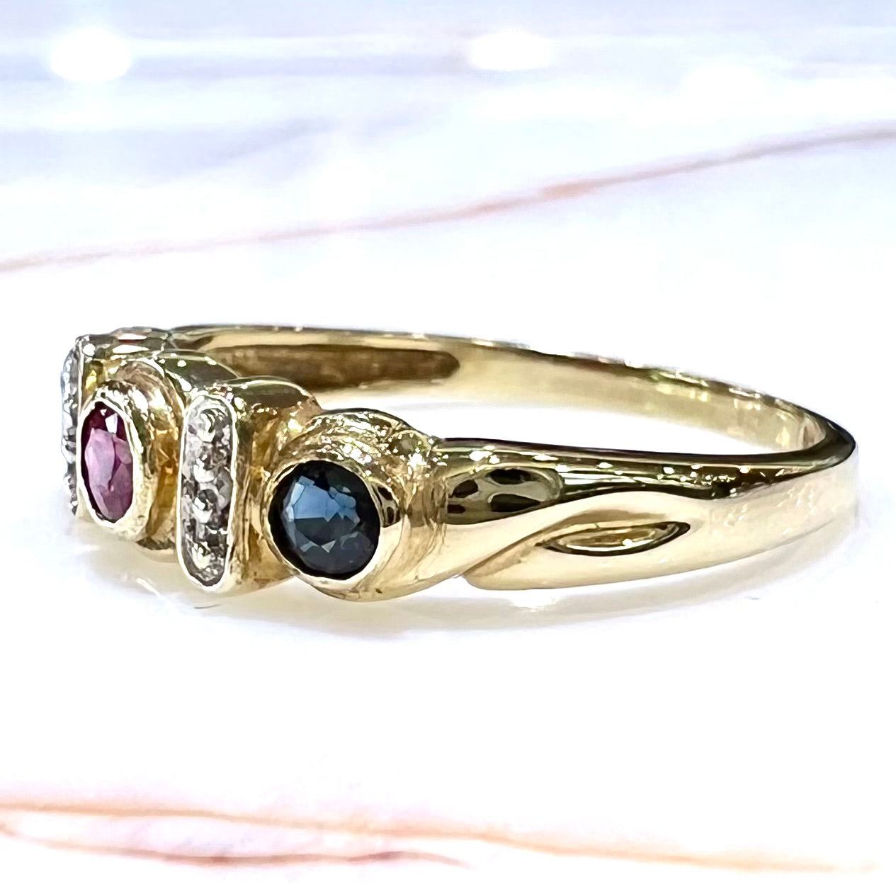 Three stone ring set with blue sapphire, lab created ruby, emerald, and two small diamonds in yellow gold.