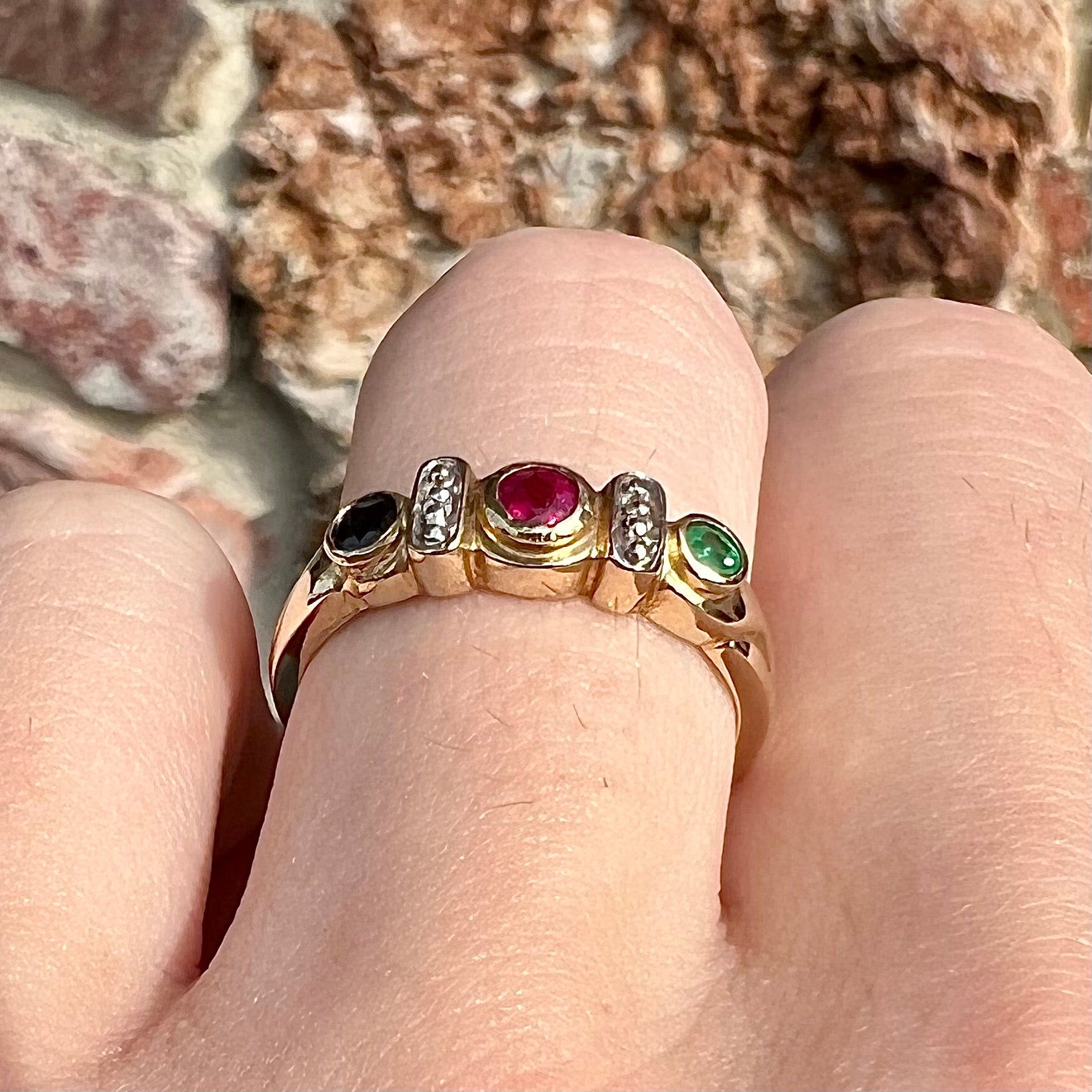 Three stone ring set with blue sapphire, lab created ruby, emerald, and two small diamonds in yellow gold.