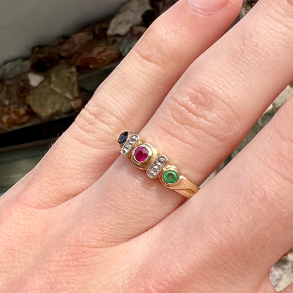 Three stone ring set with blue sapphire, lab created ruby, emerald, and two small diamonds in yellow gold.