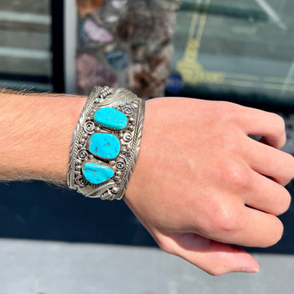 An ornate men's sterling silver turquoise cuff bracelet.  There are three turquoise stones set with silver feathers.